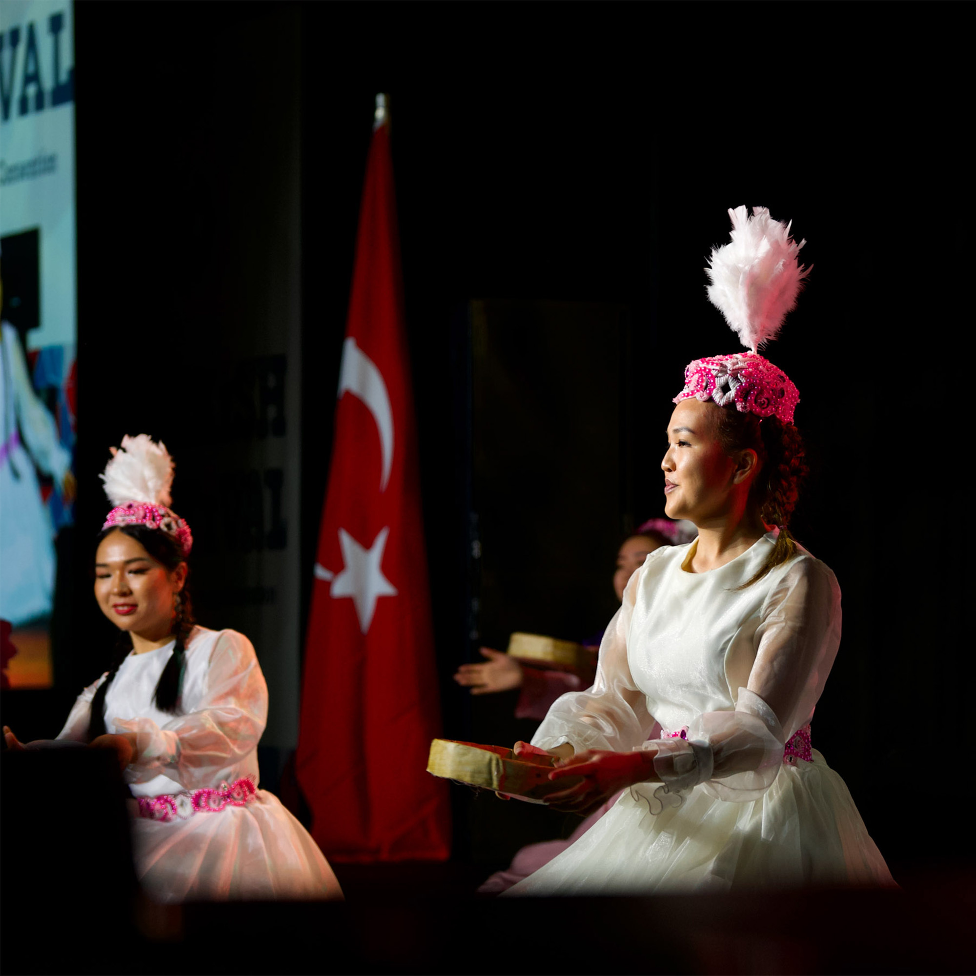 Female dancers in white, long-sleeved dresses with pink and white trim are performing on a stage. They are wearing feathery headdresses and holding small items. A large Turkish flag is seen in the background.