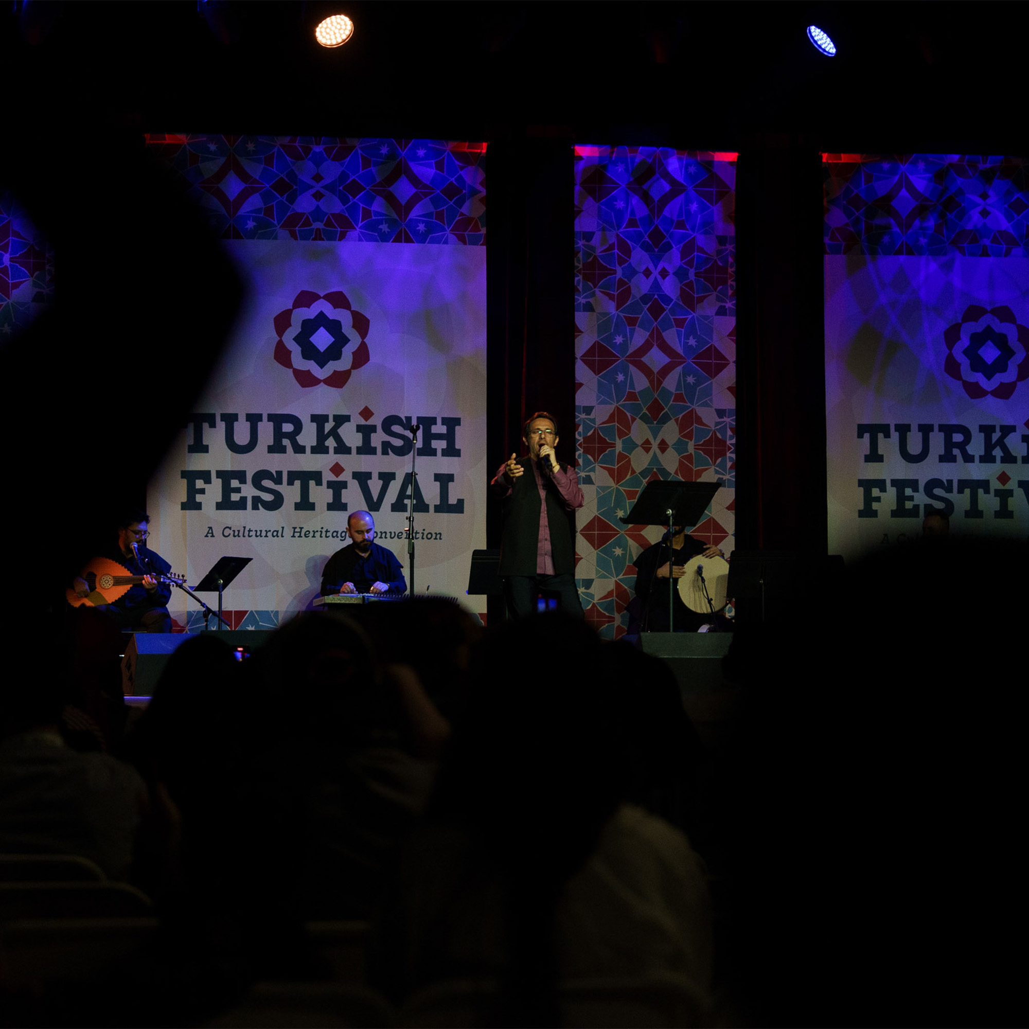 A person is performing on a stage in front of a live audience. The performer, a man, is singing into a microphone and is surrounded by musicians and their instruments. The backdrop features banners for the "TURKISH FESTIVAL" and geometric patterns. The audience's silhouettes are visible in the foreground.