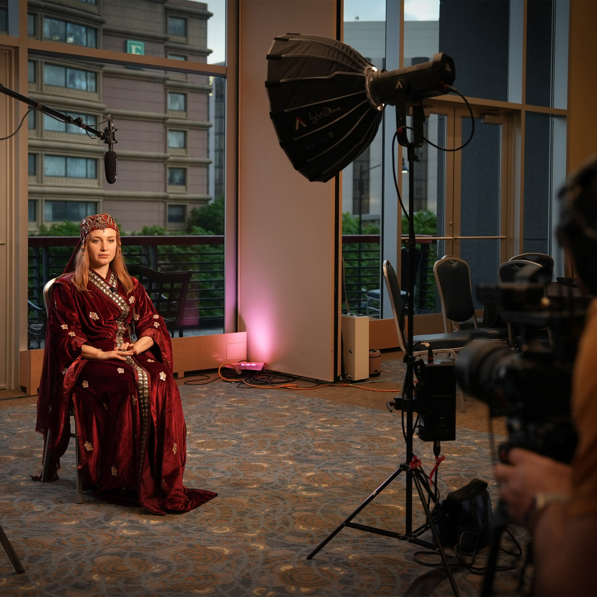 A professional video shoot is taking place in a room with a large window and a view of city buildings. A woman in elaborate traditional clothing, a red velvet dress, and a jeweled headdress is seated, facing the camera. A large light stand and a boom microphone are visible, along with a camera filming her.