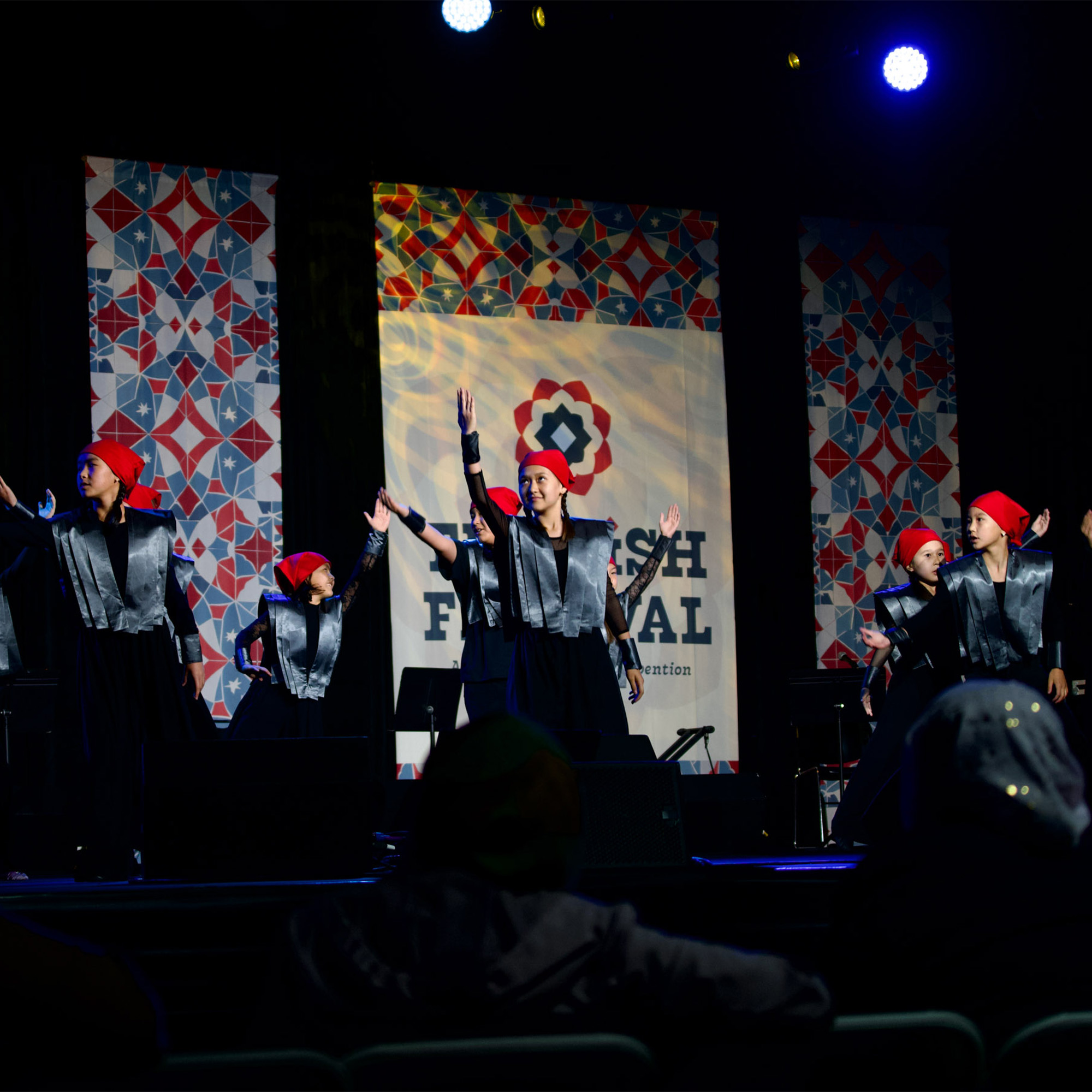 A performance is taking place on a stage with a dark backdrop and large screens displaying colorful, geometric patterns. A group of dancers, female, are performing in matching black and silver costumes with red head wraps. They have their arms raised in unison. The stage is lit with blue and white spotlights, and an audience member's head is visible in the foreground.