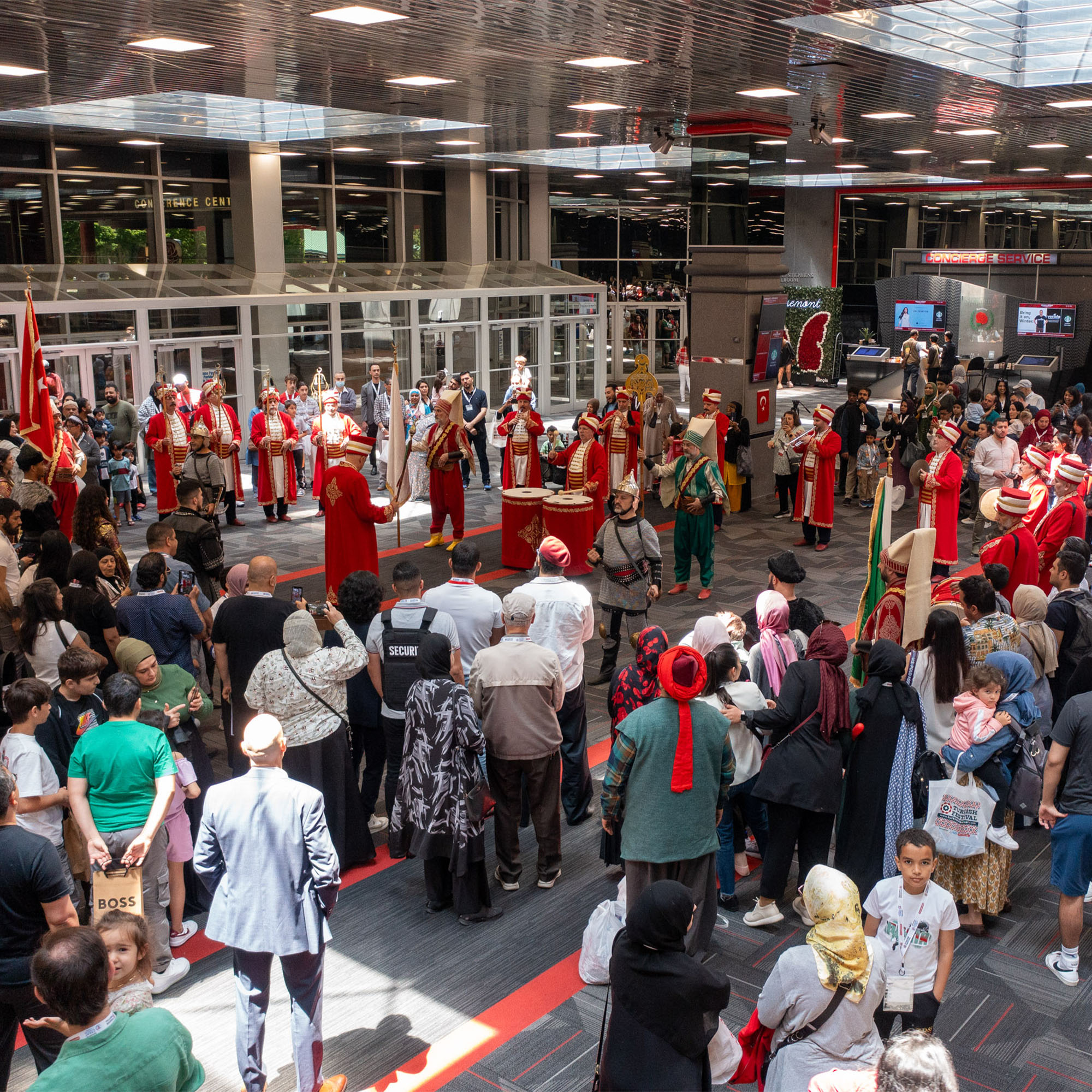 A wide-angle shot from a high perspective shows a large crowd of people gathered in a spacious indoor hall. In the center, a group of performers in elaborate red and green traditional costumes are performing with drums and flags. The crowd is watching the performance, and the setting is an cultural event.