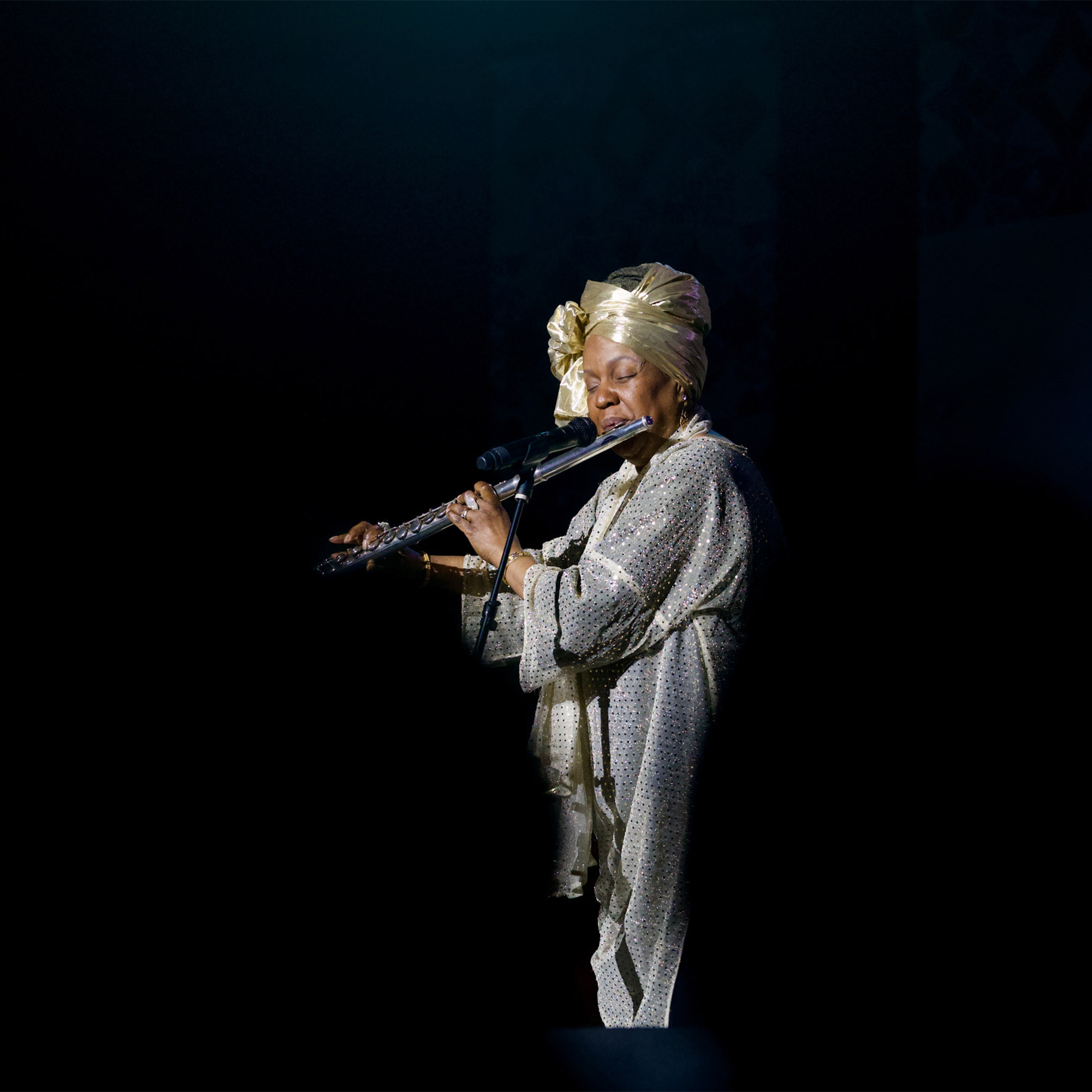A woman is on a dark stage, playing a silver flute. She is wearing a shimmering gold and silver dress and a matching head wrap. A microphone is positioned in front of the flute to capture the sound. She is illuminated by a spotlight, with the rest of the stage in darkness.
