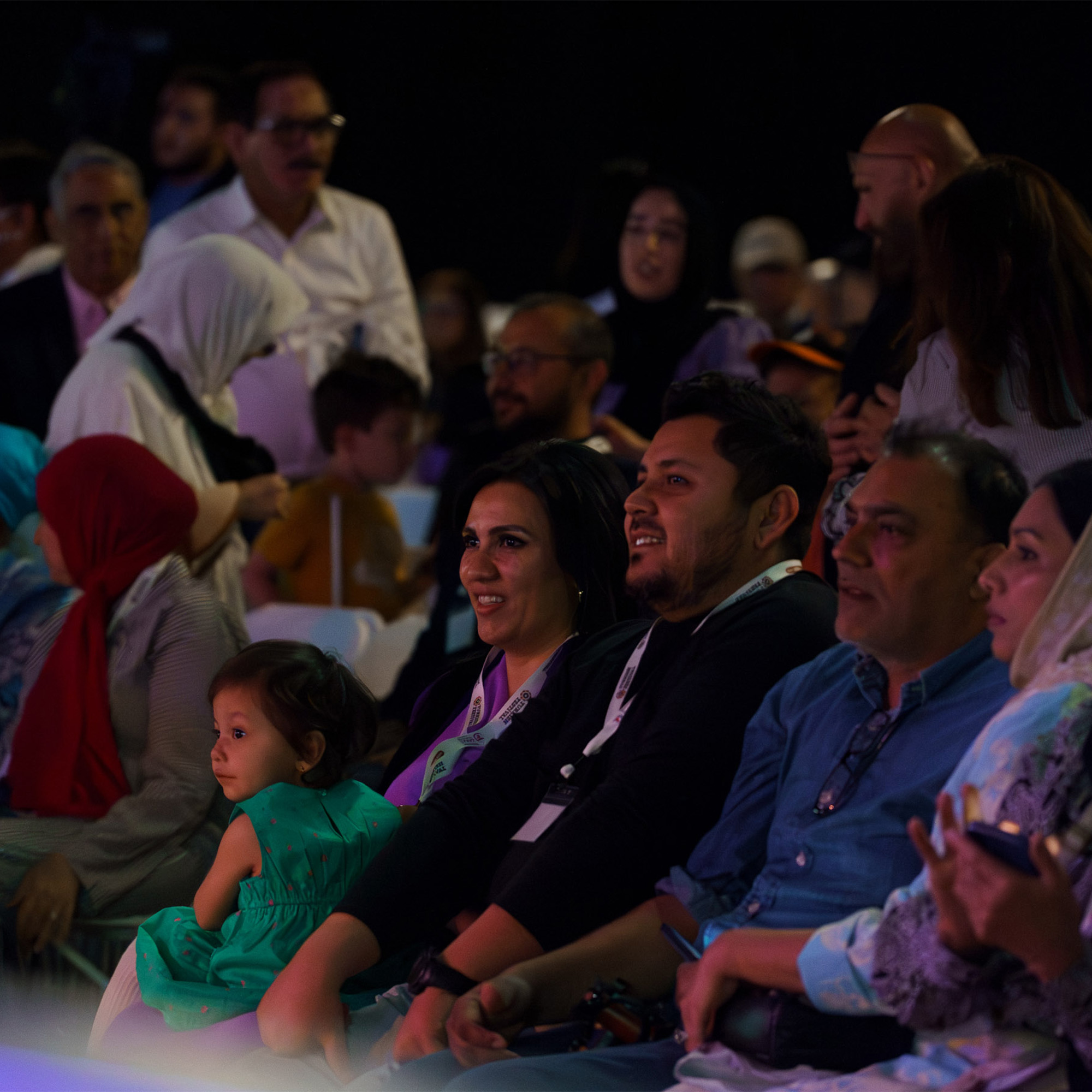 A candid shot of an audience at an event. A group of people are seated and looking toward a performance, with many of them smiling. A young child is seated in the front row. The audience is diverse, with men, women, and children. The lighting is low.
