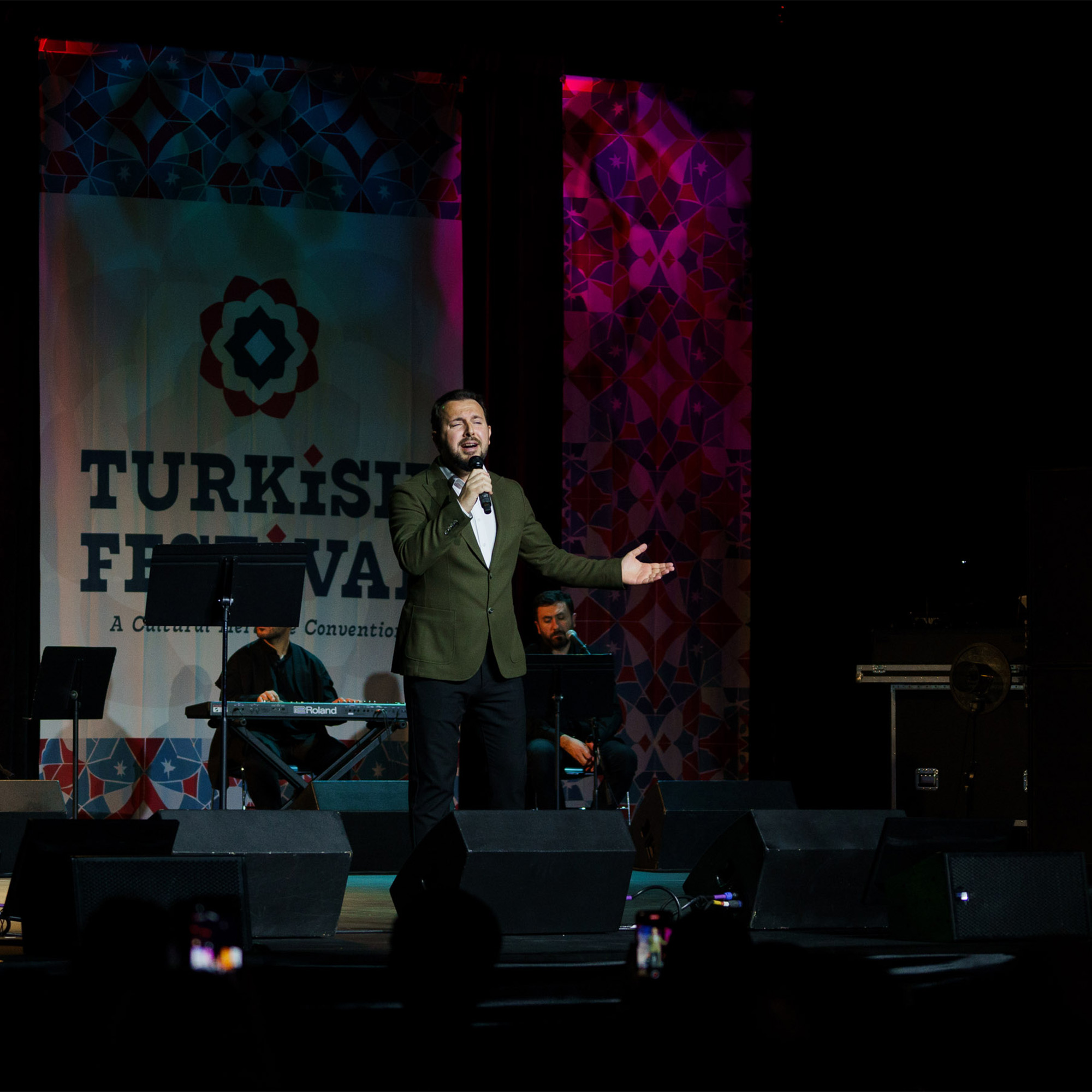 A man in a green jacket and a white bow tie is performing on a stage, holding a microphone and gesturing with his left hand. In the background, a large screen displays a banner for the "TURKISH FESTIVAL" and a musician is seated at a keyboard. The stage is lit with colored lights.
