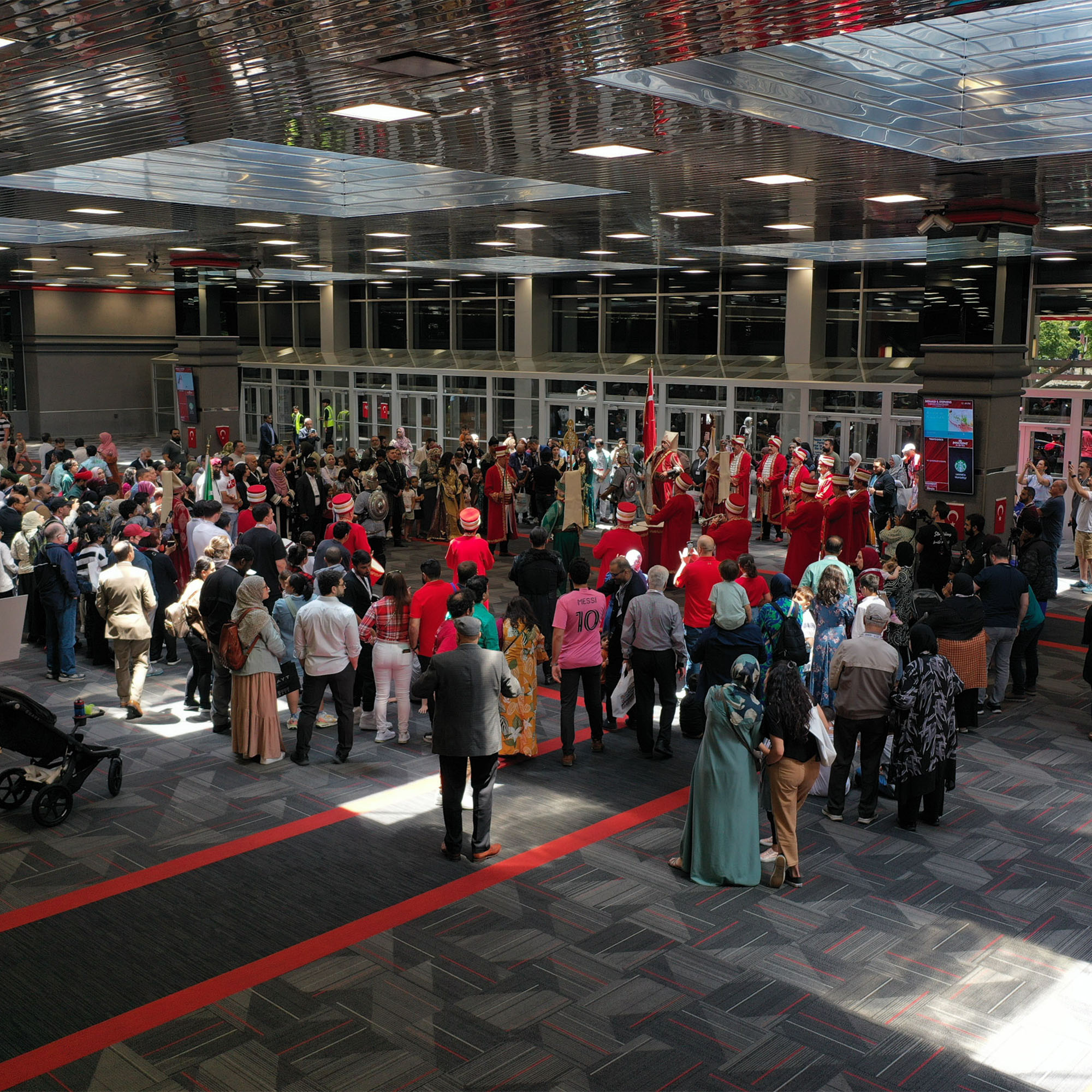 A wide-angle shot from a high perspective shows a large crowd of people gathered in a spacious indoor hall with a checkered floor and a modern ceiling. In the center of the crowd, a group of performers in what appear to be traditional wear, primarily in red and white, are performing.