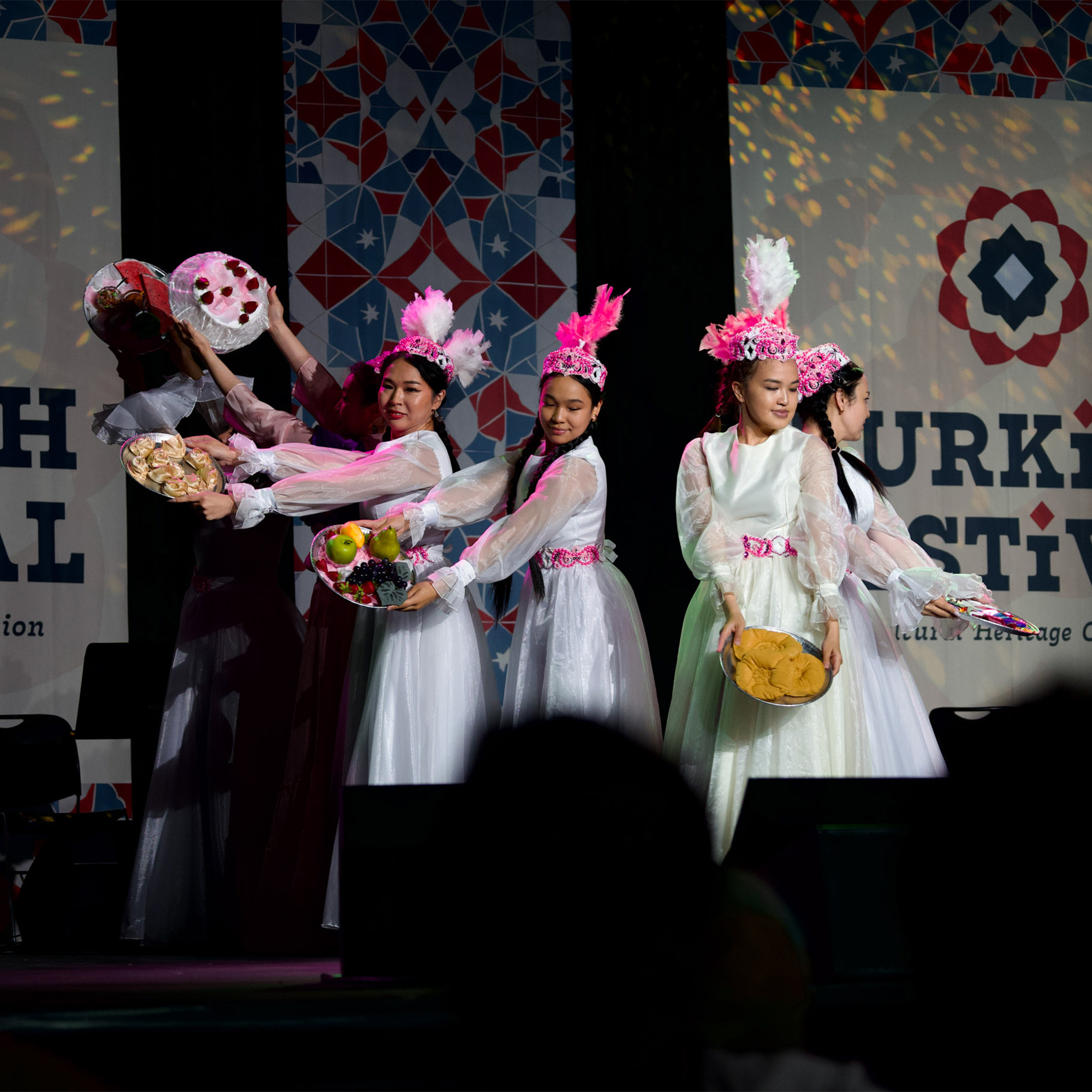 A group of four female dancers are on a stage, performing in traditional costumes. They are wearing white dresses with pink and white hats, and are holding trays of food. They are performing against a backdrop with a repeating pattern and a banner for the "TURKISH FESTIVAL."