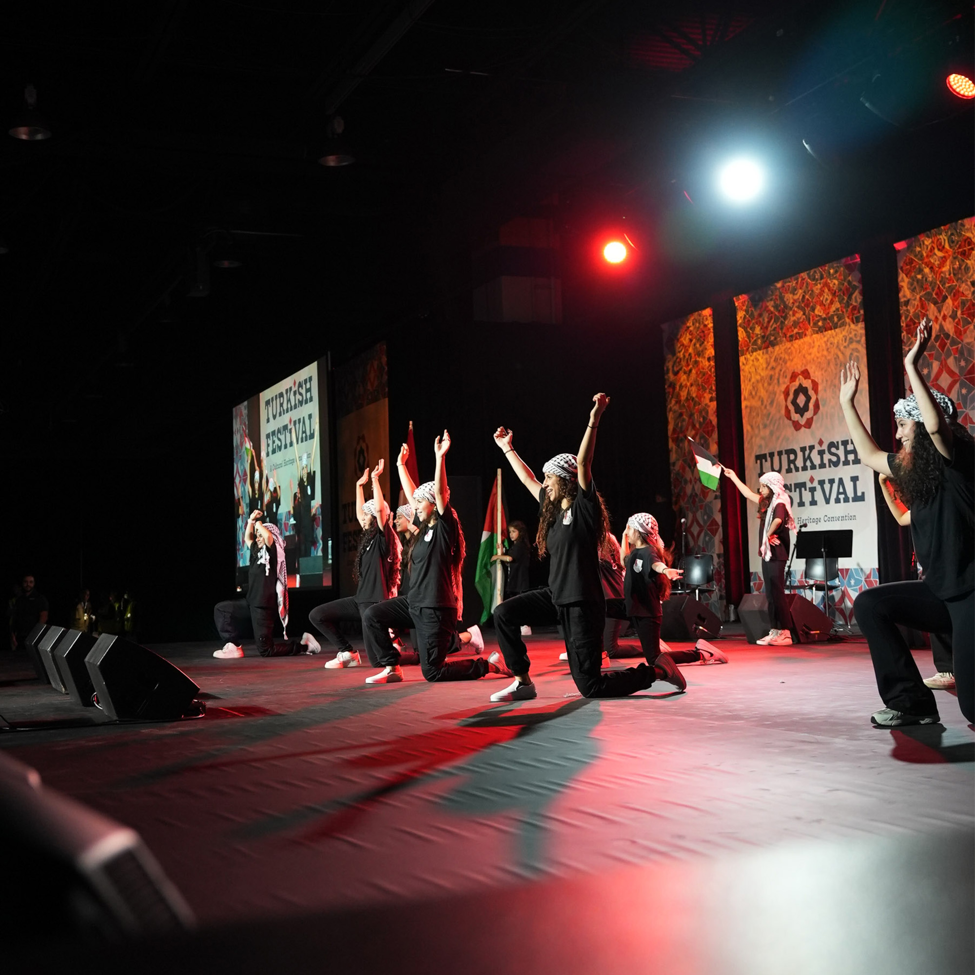 A shot of a stage shows a group of performers, both men and women, kneeling with their arms in the air, wearing black t-shirts and head coverings, and are performing under a red and white spotlight. Large screens in the background display "TURKISH FESTIVAL" and colorful patterns. A Palestine flag is also visible on the stage.