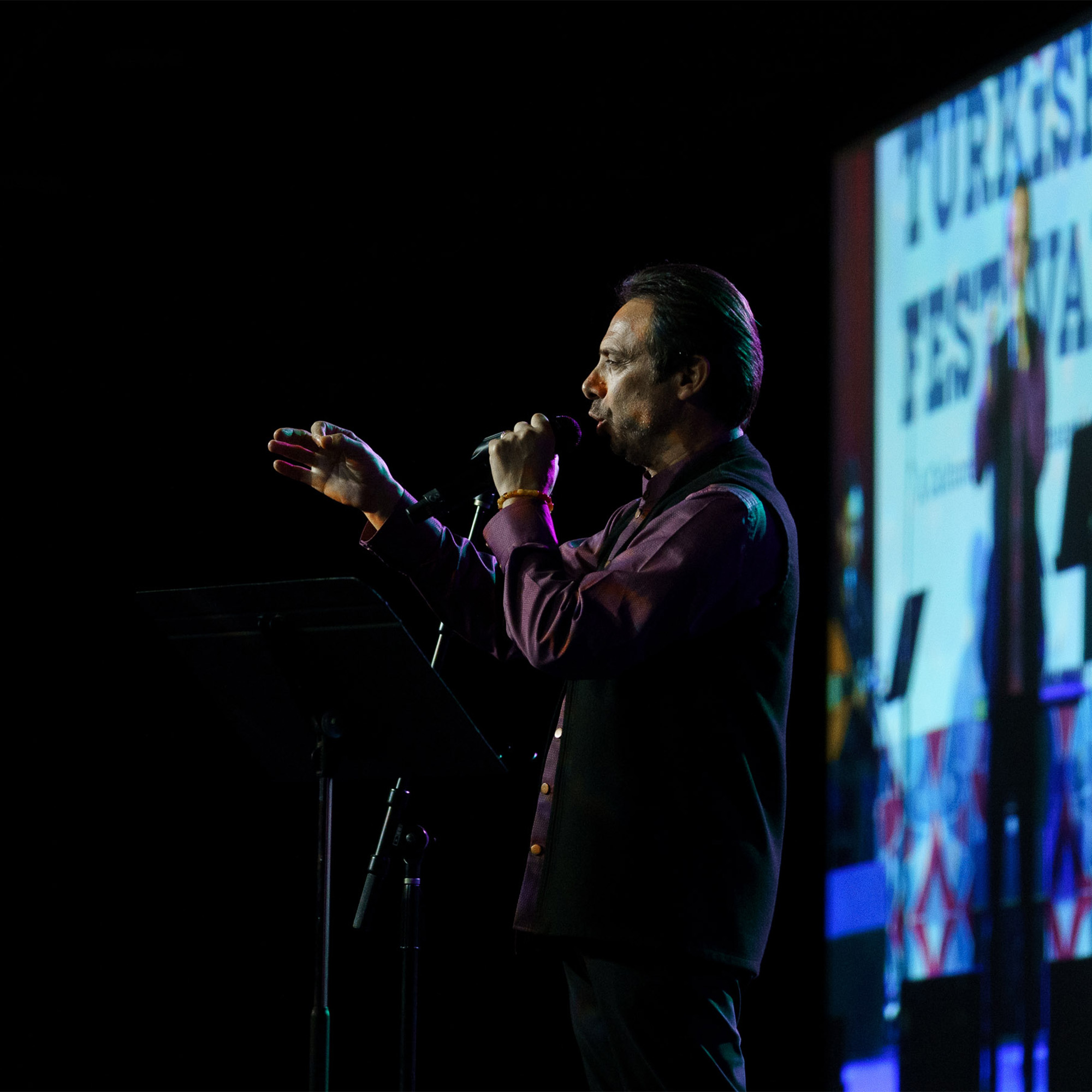 A side profile shot shows a man on a stage, singing into a microphone and gesturing with his hands. He is wearing a dark vest over a purple shirt. A large screen in the background shows the words "TURKISH FESTIVAL," and the stage is lit with colored lights.