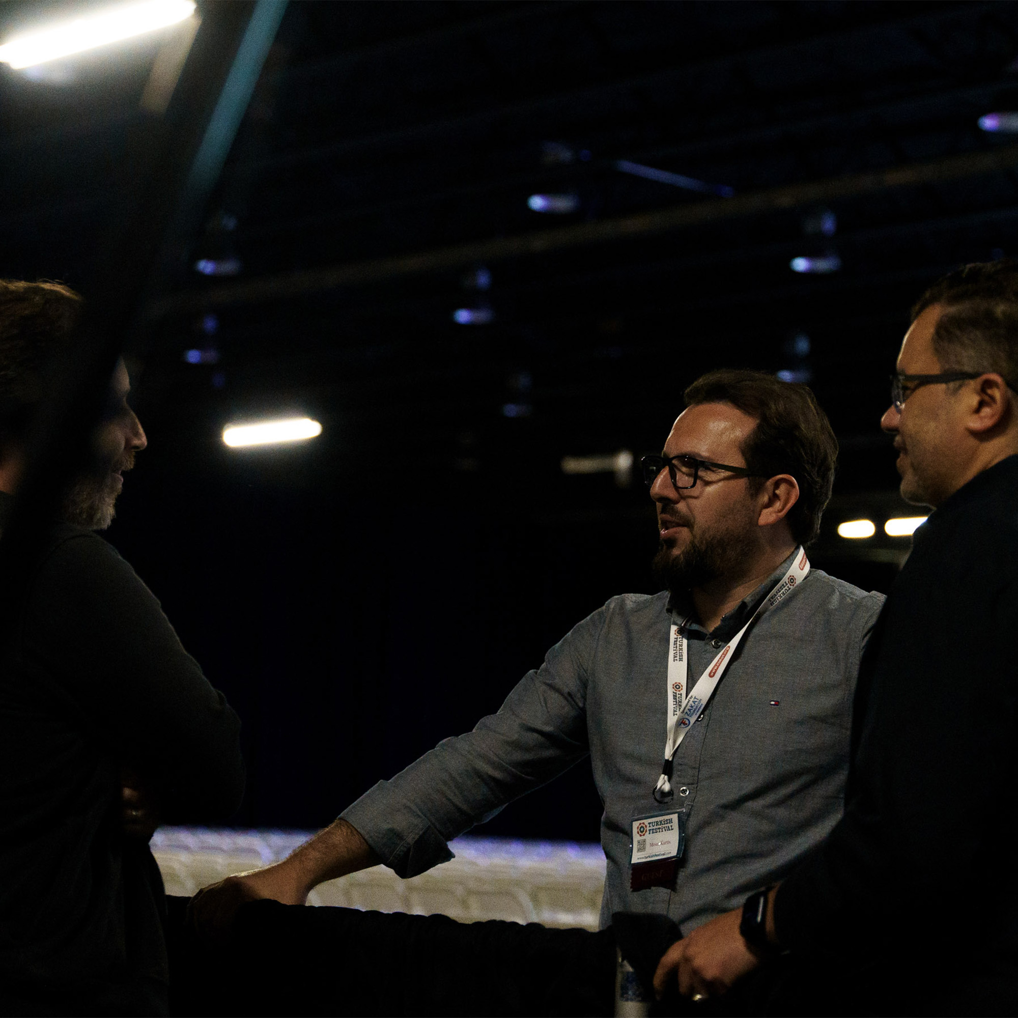 A shot shows three men in a dimly lit indoor space. They are standing and talking, with the man in the center looking at the man on the right. The man in the center is wearing a gray shirt and glasses, and has an event badge hanging around his neck.